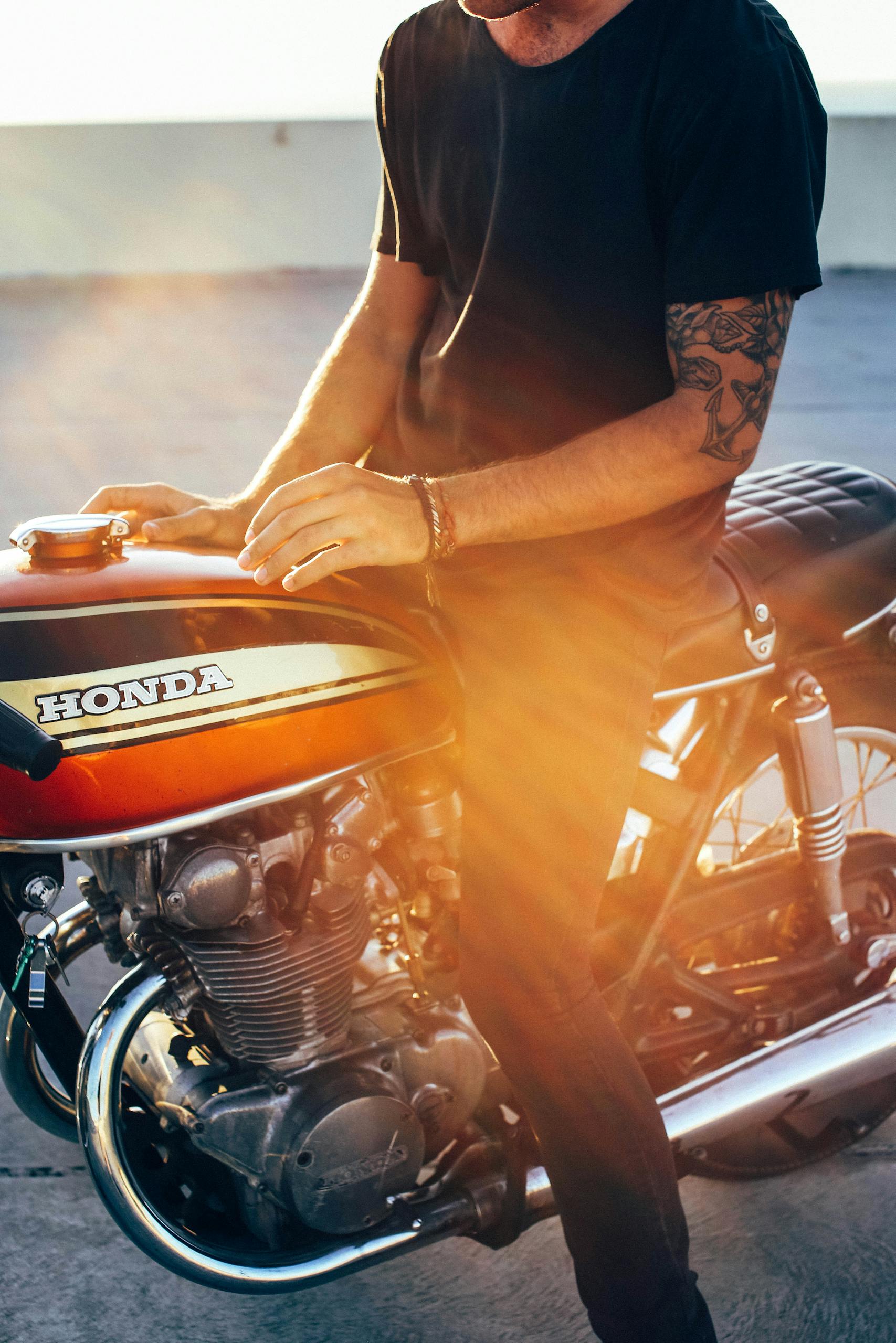 Crop anonymous young tattooed male biker in casual clothes sitting on modern motorcycle on asphalt road on sunny day